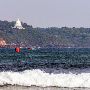 A photograph of Rumassala Hill in Sri Lanka, showing a lush, green hill with an ancient-looking banyan tree, overlooking the calm, blue Indian Ocean and the distant Galle Fort at sunrise.